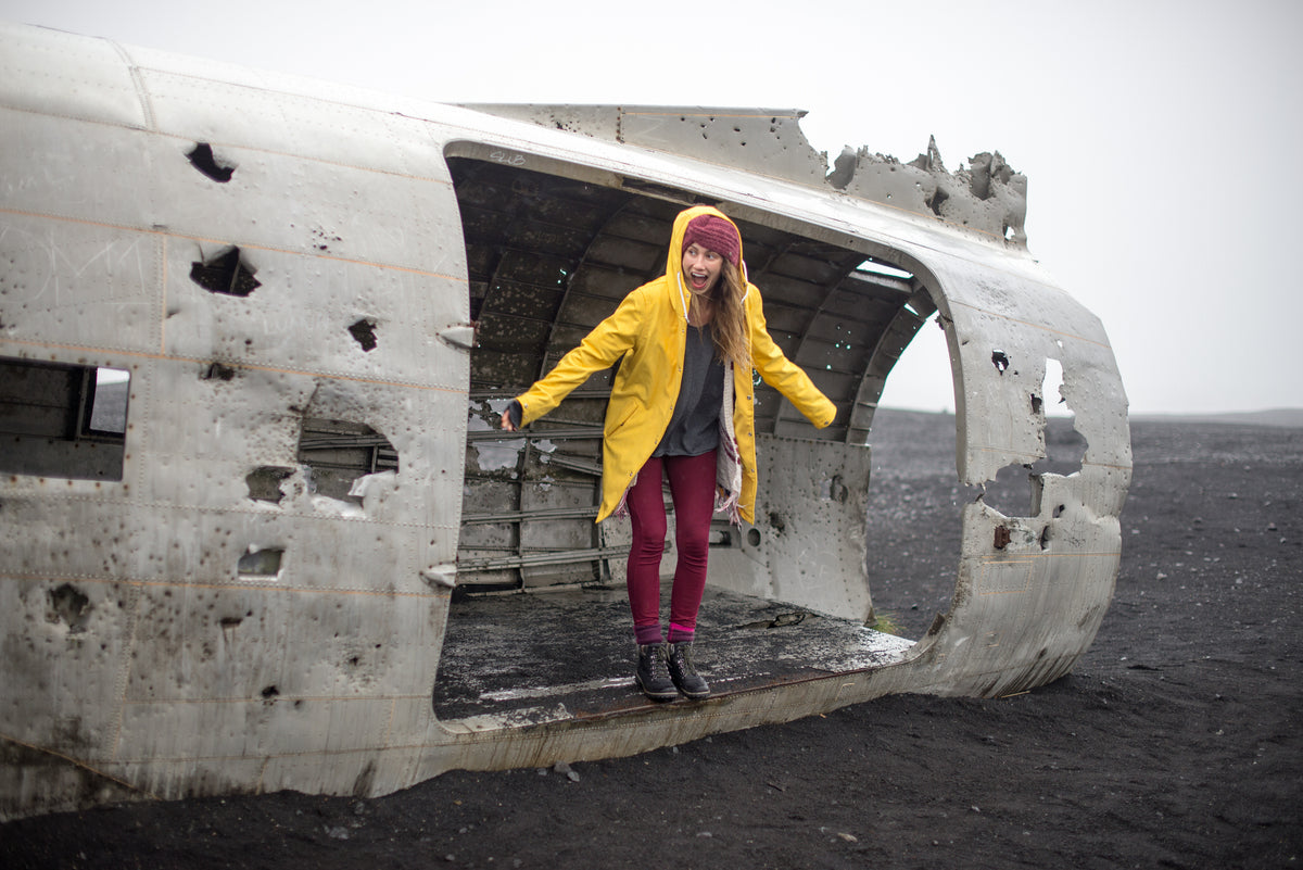 Woman in the yellow Original raincoat stepping out of an old plane wreck on black sand