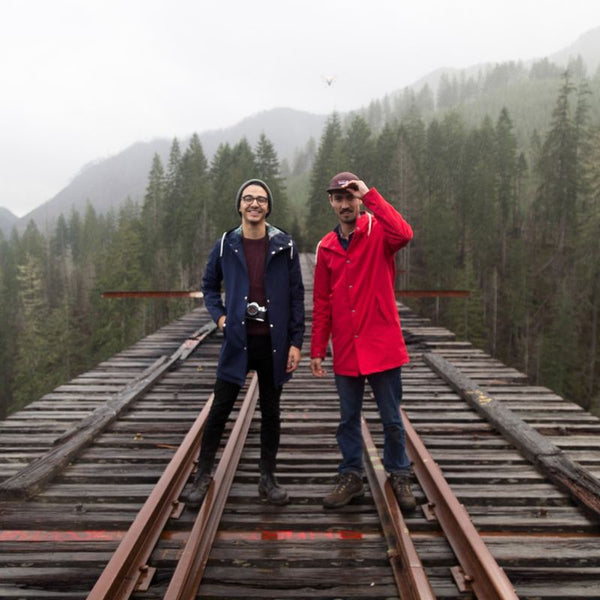 Two people in Ember & Earth rain jackets on a forest railway bridge