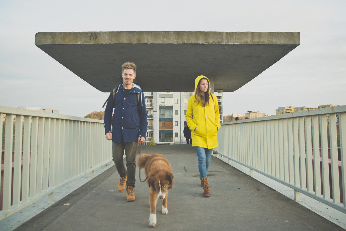 Couple walking a dog across a bridge, one wearing the Ember & Earth Yellow | The Original rain jacket